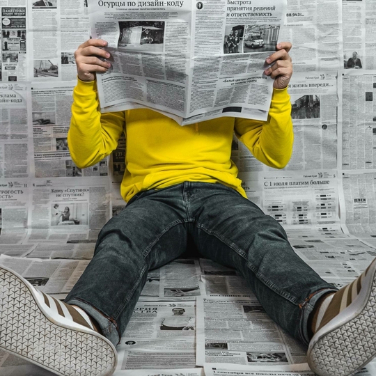 Man reading newspaper while sitting on the floor in a room covered in newspapers.